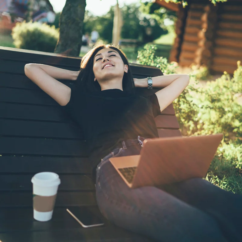 A woman leans back with closed eyes on an outdoor bench. She has finished working on her laptop and is enjoying her life after treatment at Sheridan Grove Recovery.