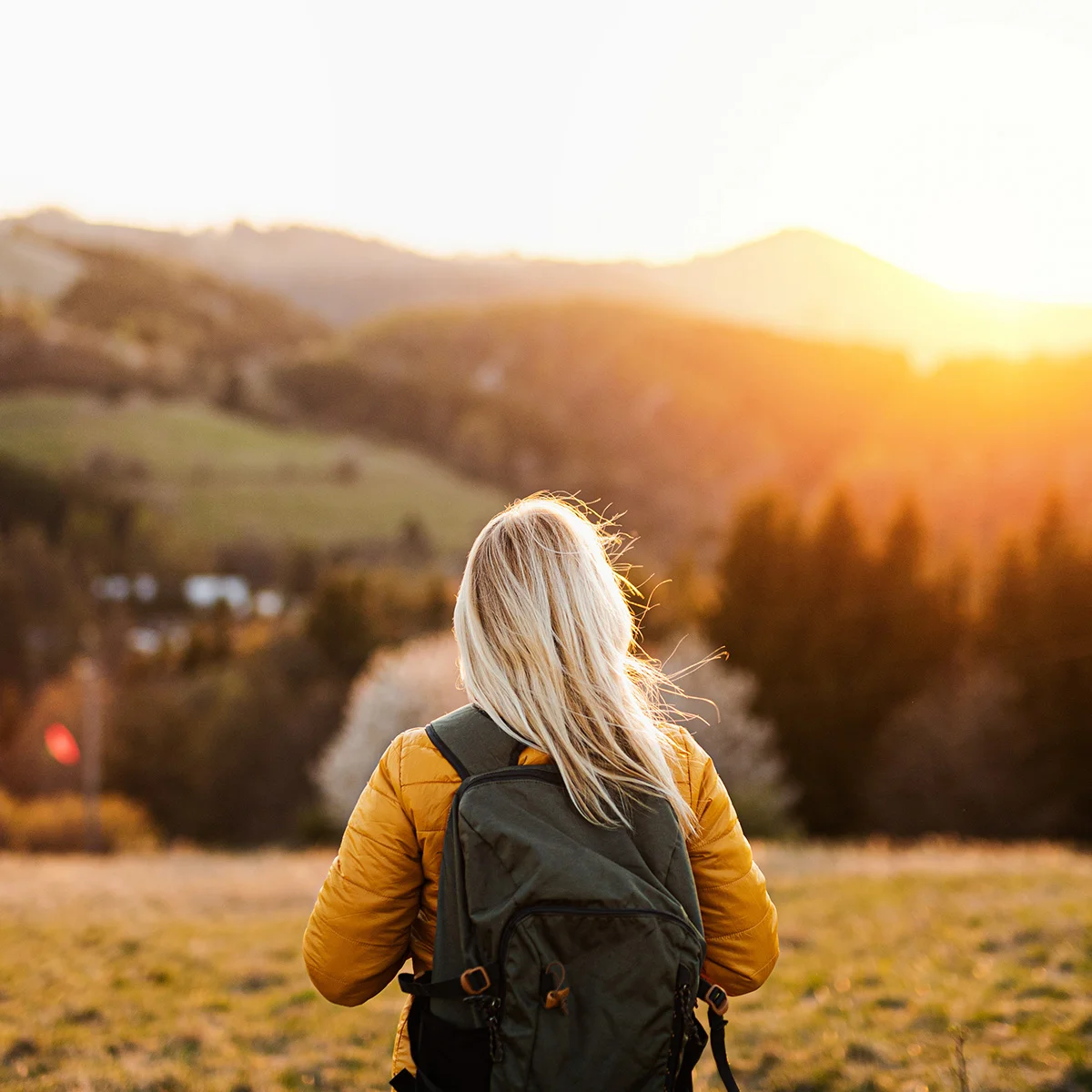 A woman wearing a backpack, stands with her back to the camera. The mountains before her are lit by golden hour. She is happy to have been able to pay for addiction treatment.
