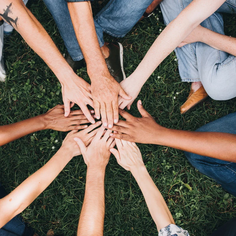 A group of people sit cross-legged on the grass with their hands extended toward one another. They are making a pact to support one another in aftercare at Sheridan Grove Recovery.